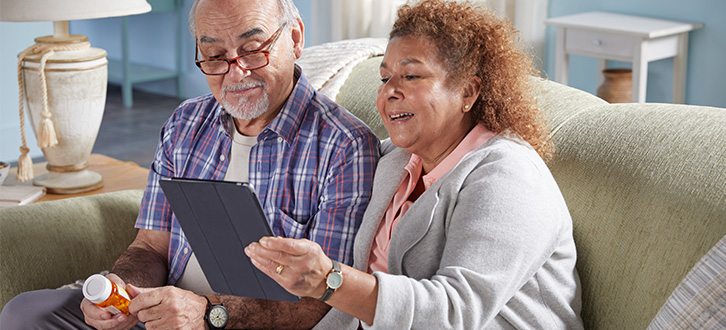 Man and woman sitting in living room smiling, reviewing their tablet device with prescription in hand.​​ 